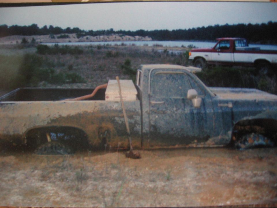Mud-caked blue square-body Chevy pickup sunk to the rockers at a sand pit