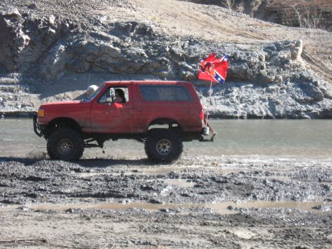 1987 Ford Bronco - Red 1987 Ford Bronco navigates a muddy off-road course near a body of water.