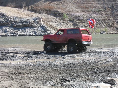 1987 Ford Bronco - This lifted SUV is navigating a muddy, watery terrain, with a Confederate flag visible on a pole in the distance.