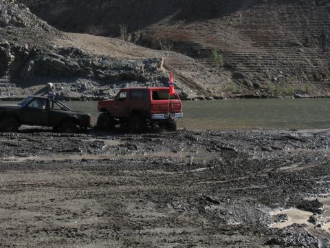 1987 Ford Bronco - Two off-road vehicles are stuck in thick mud next to a body of water, with a rocky hillside in the background.