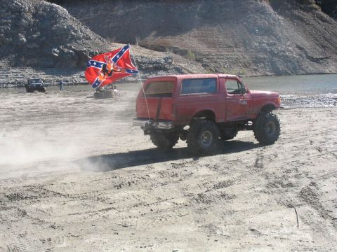 1987 Ford Bronco - A red 1987 Ford Bronco is shown lifted with large tires, kicking up dust near a body of water, with a Confederate flag flying.