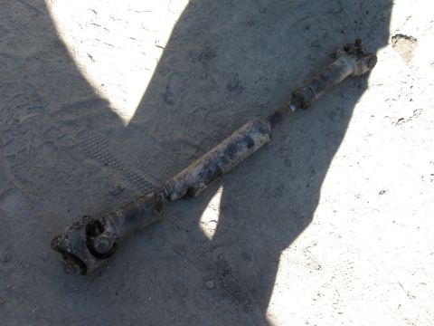 1987 Ford Bronco - A removed driveshaft lying on dusty ground in bright sun, showing the slip yoke, splined shaft and U-joints at both ends caked in dirt and…