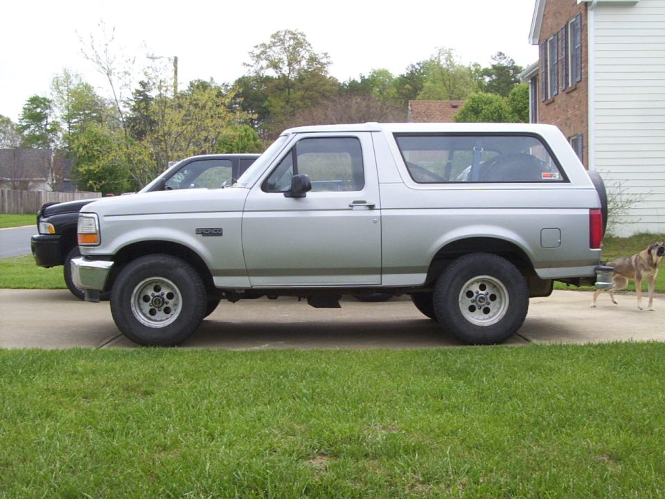 Side view of a silver Ford Bronco SUV with rear spare tire in a driveway, dog standing behind it