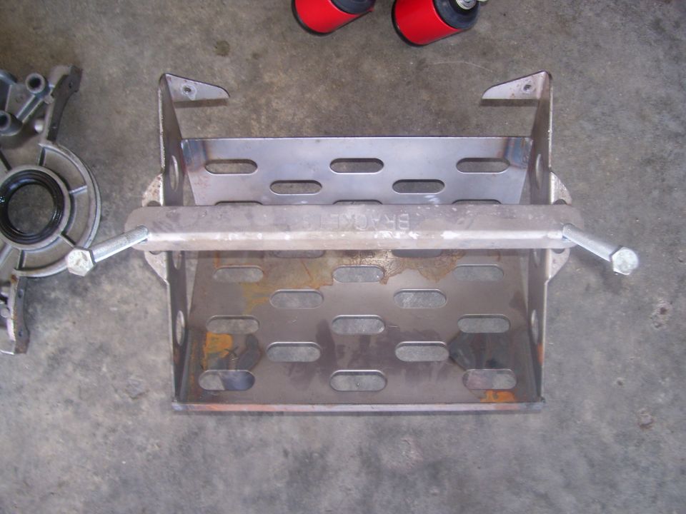Metal battery tray with slotted holes and a crossbar, next to a silver engine component