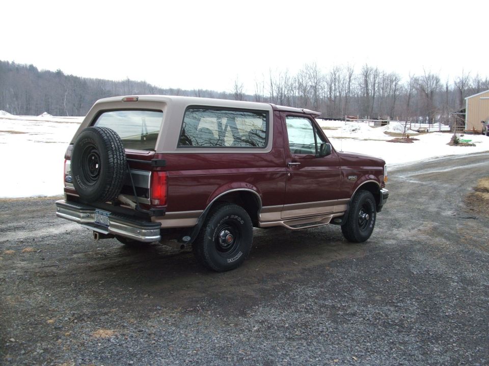 Maroon Ford Bronco SUV with tan lower trim and rear spare tire on a gravel drive with snow and trees behind