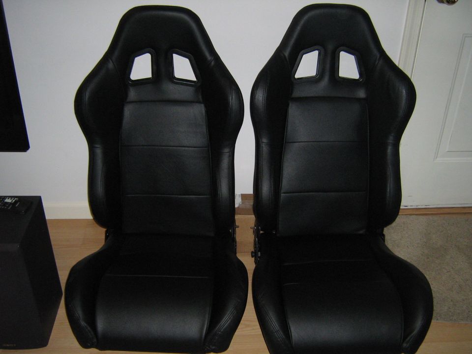 Two black racing seats with headrest cutouts and stitched panels, positioned side-by-side indoors