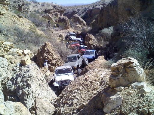 Several off-road vehicles navigate a narrow, rocky canyon. One white Jeep is in the foreground