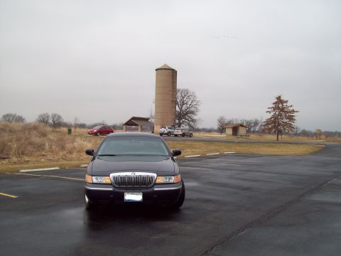 1999 Mercury Grand Marquis - A dark sedan is parked in a lot with a silo and buildings in the distance.