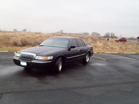 1999 Mercury Grand Marquis - Dark sedan parked in a lot with dry grass and overcast sky. Another car is visible in the distance.