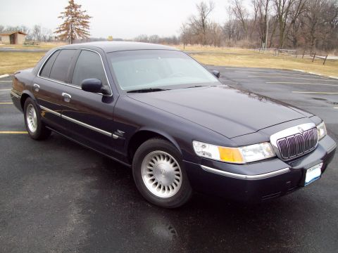 1999 Mercury Grand Marquis - A dark blue sedan is parked on a wet asphalt surface with trees in the background.