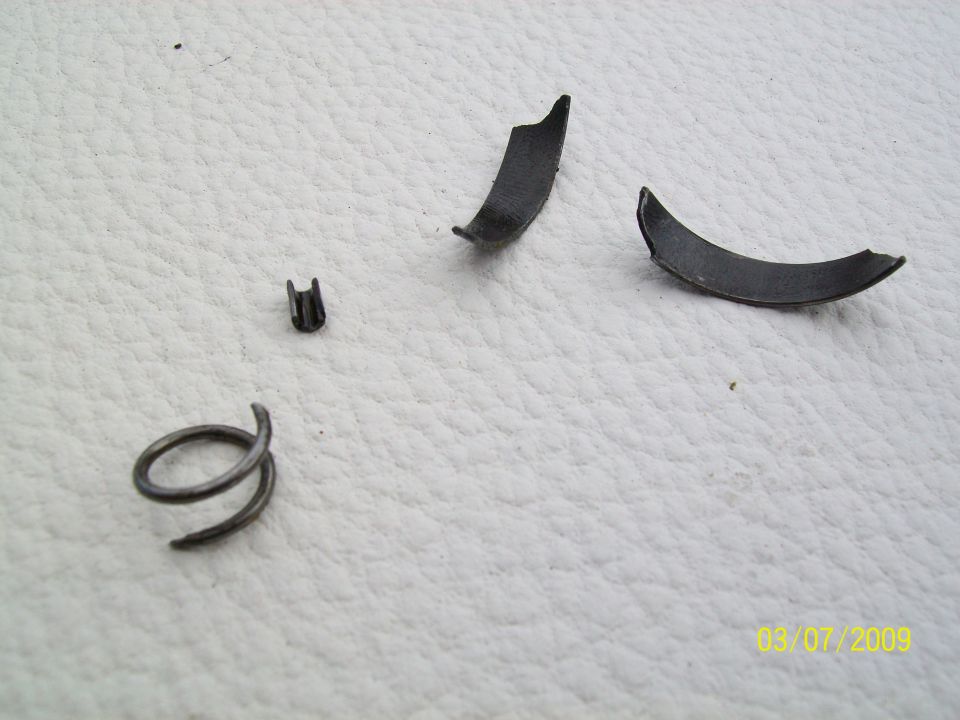 Broken spring, clip, and curved metal pieces on a textured surface. Watermark reads 03/07/2009 SUPERMOTORS.NET