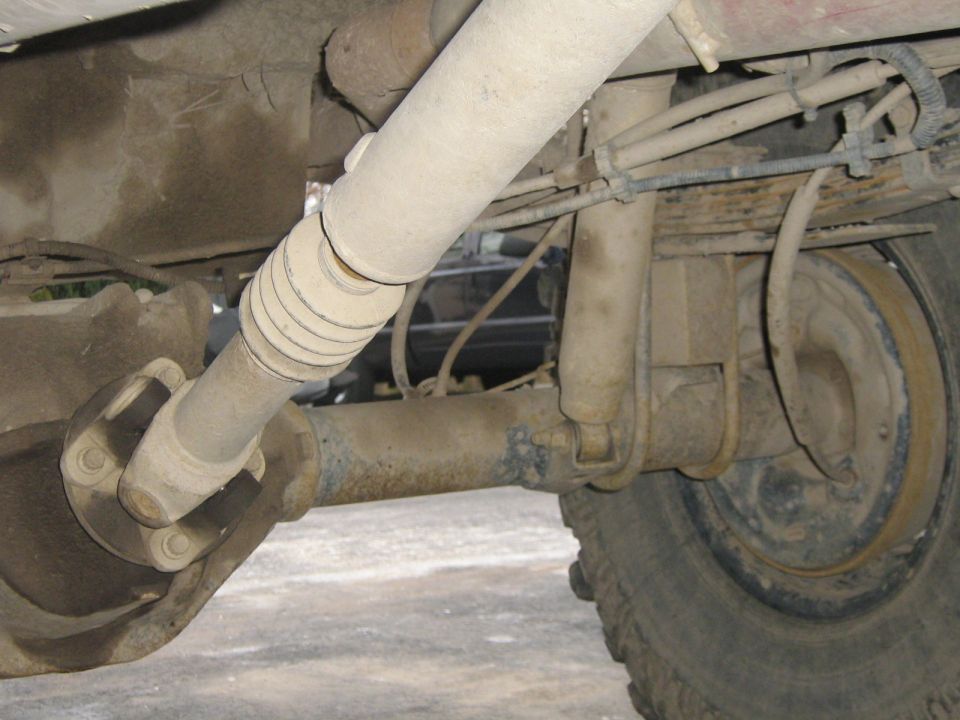 Underside view of a dusty rear axle, driveshaft, leaf springs, and shock absorber on a vehicle