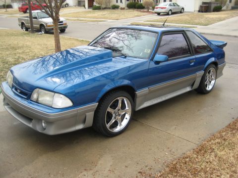1990 Ford Mustang - A blue Ford Mustang with a prominent hood scoop sits on a concrete driveway in a residential neighborhood.