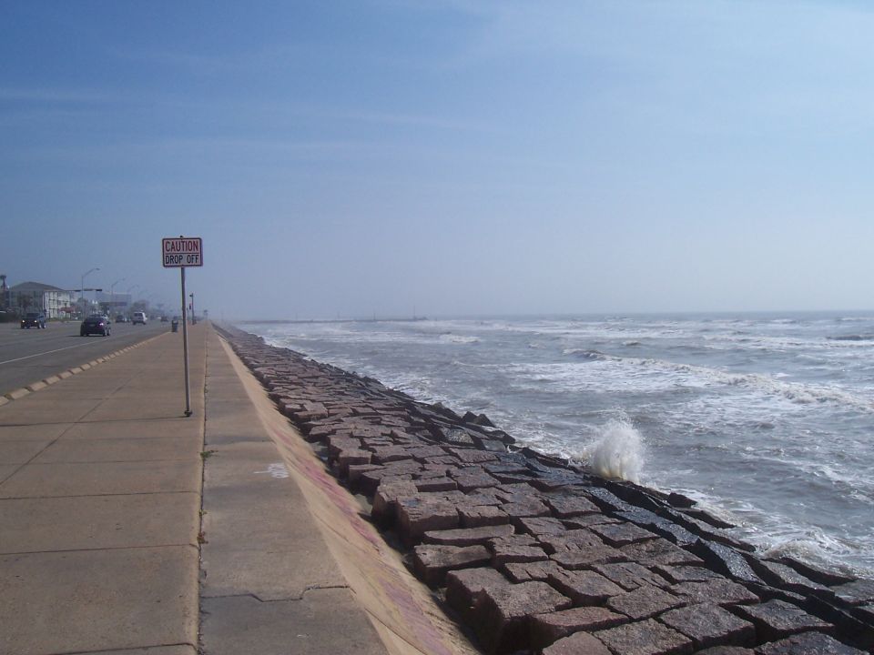road next to a stone sea wall with waves crashing, caution drop off sign visible