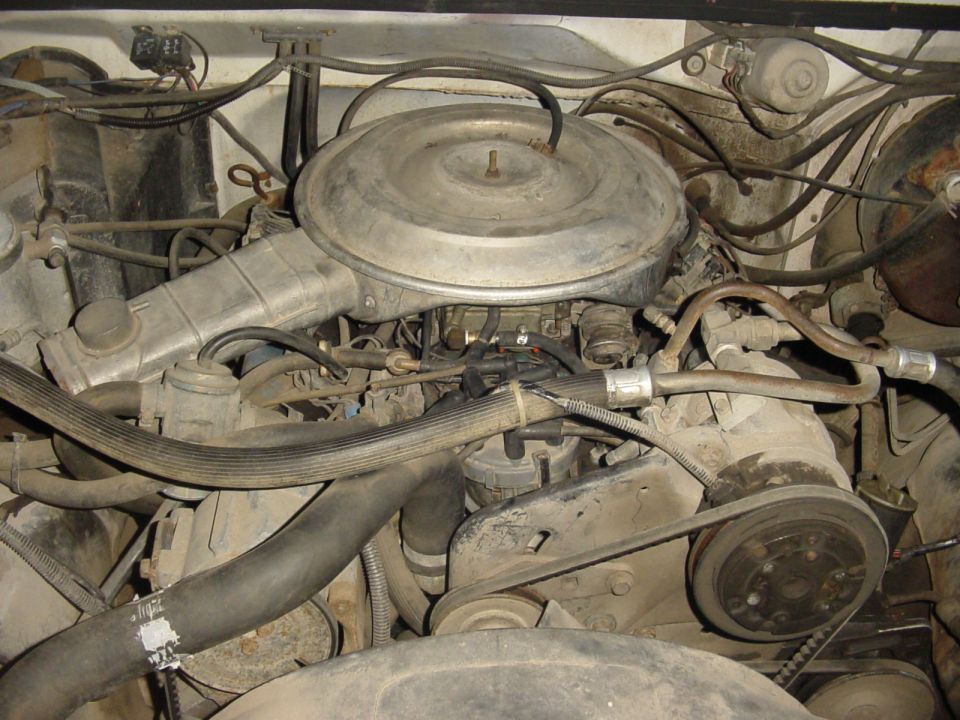 Dusty engine bay with round air cleaner, carbureted V8, belts, pulleys and radiator hose