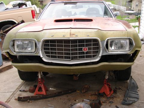 1973 Ford Ranchero - A faded green Ford project car with a hood scoop, spelled-out FORD on the nose and a honeycomb grille sits on jack stands in a yard, front…