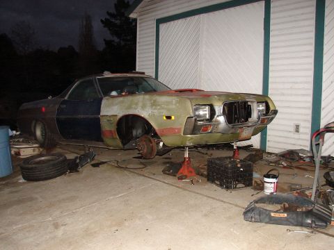 1973 Ford Ranchero - A 1970s Ford Ranchero car-pickup mid-restoration sits on jack stands in a driveway at dusk, front-left brake hub bare with the wheel remove…