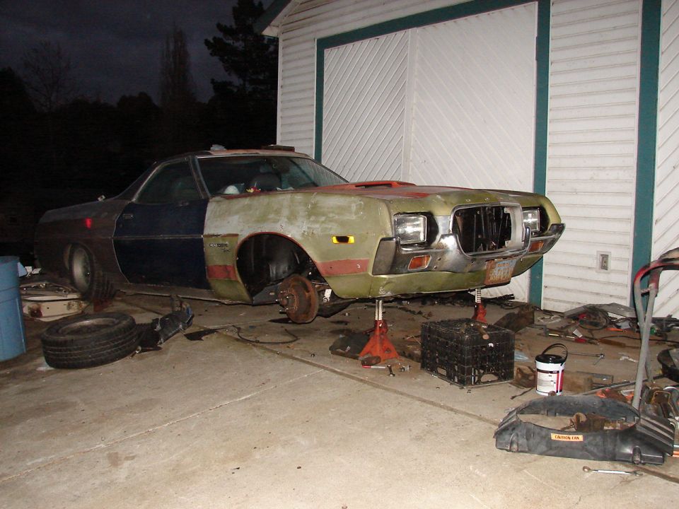 Weathered green primer 1970s Ford Ranchero on jack stands with front wheel off, front hub exposed, in a driveway
