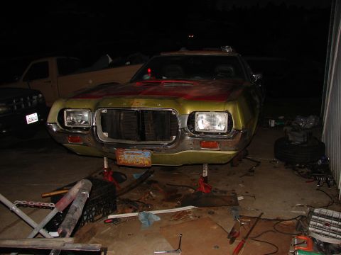 1973 Ford Ranchero - Night shot of a green Ford project car sitting on jack stands on a concrete driveway, front end stripped with the grille removed and FORD l…