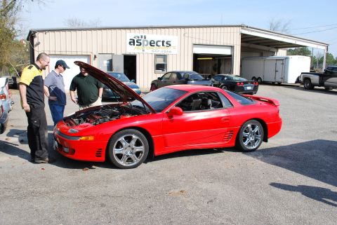 1991 Mitsubishi 3000GT - Three men examine the engine of a red 1991 Mitsubishi 3000GT parked outside a commercial building.