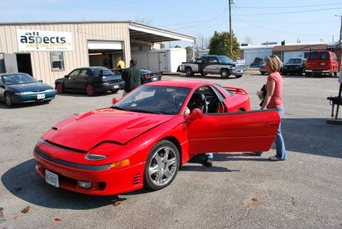 1991 Mitsubishi 3000GT - A red 1991 Mitsubishi 3000GT is parked outside a commercial building with its door ajar.