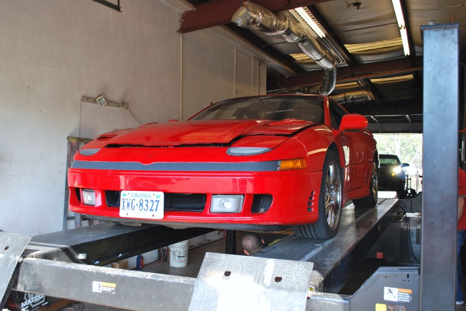 Red 1991 Mitsubishi 3000GT on a car lift, Virginia license plate XWG-8327 visible. Another car is in the background