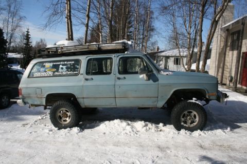 1990 Chevrolet Suburban - A light blue, lifted Chevrolet Suburban is parked in the snow with a roof rack loaded with gear. The vehicle has several stickers on the re…