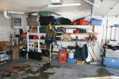 1990 Chevrolet Suburban - A cluttered garage scene with shelves full of storage bins, tools, and various items. The concrete floor is wet in several areas, suggestin…