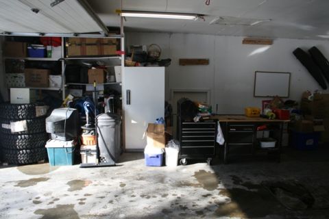 1990 Chevrolet Suburban - This is a cluttered garage scene with shelves full of boxes, tires, a television, and a trash can. A workbench with tools is visible on the…
