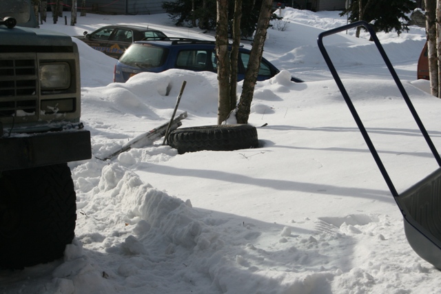 Dark SUV and truck buried in snow, with a tire and shovel visible.