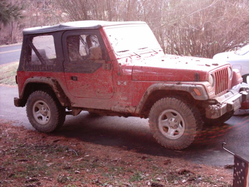 Red Jeep Wrangler with black soft top, covered in mud, parked on the side of the road.