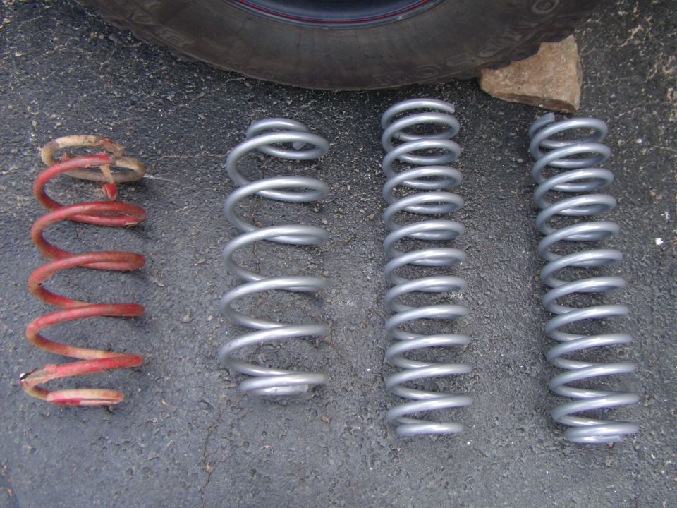 Four coil springs on asphalt: one red and worn, three new silver.