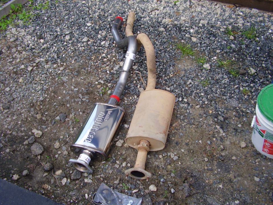 Two exhaust mufflers, one chrome and one rusted, on a gravel surface.