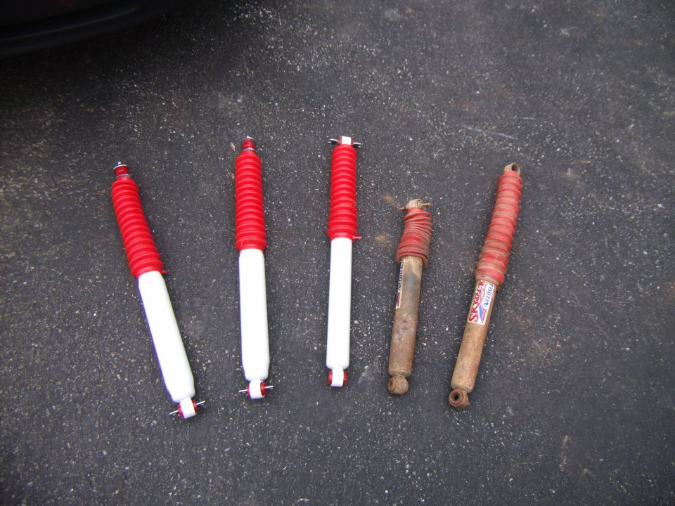 Five shock absorbers on asphalt, three white and red, two rusty metal and red.