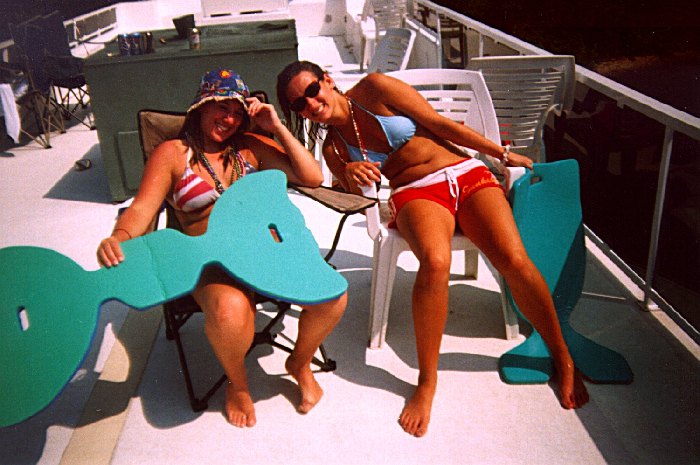 Two women in swimwear with teal foam floats on a houseboat's white upper deck