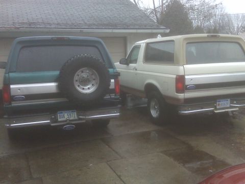 1995 Ford Bronco - The rear view of two Ford Broncos parked on a wet concrete driveway. The Bronco on the left is dark green with a spare tire mounted on the…