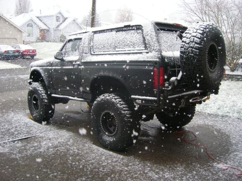 1996 Ford Bronco - Heavily lifted flat-black Ford Bronco dusted with snow in a suburban driveway during falling snow, seen from the driver-side rear three-qua…
