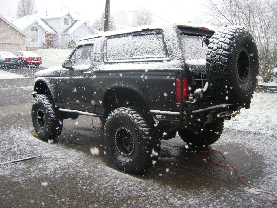 Lifted flat black Ford Bronco on mud tires with rear swing-out spare, covered in falling snow