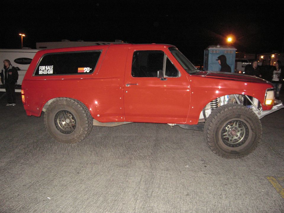 Red Ford Bronco with off-road tires and suspension mods, side view at night.
