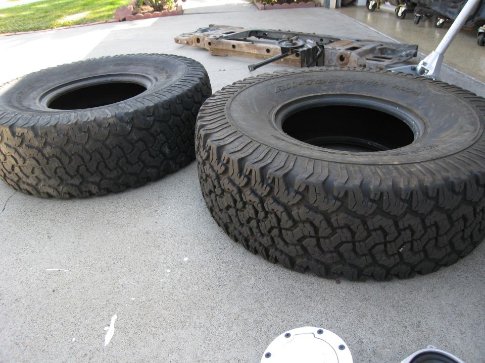 Two all-terrain tires on a concrete surface with a vehicle frame in the background.