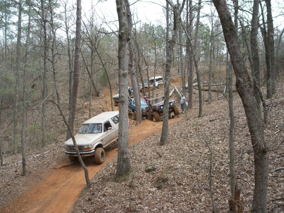Tan 1996 Ford Bronco driving up a wooded dirt trail with other off-road vehicles.