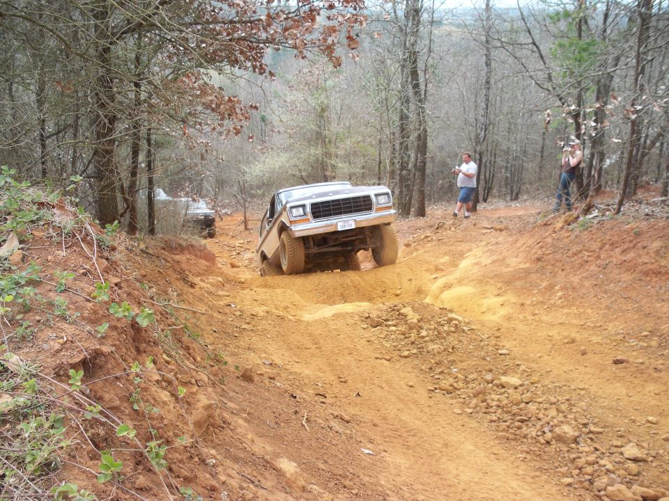 Tan Ford Bronco driving up a steep dirt trail in a forest.