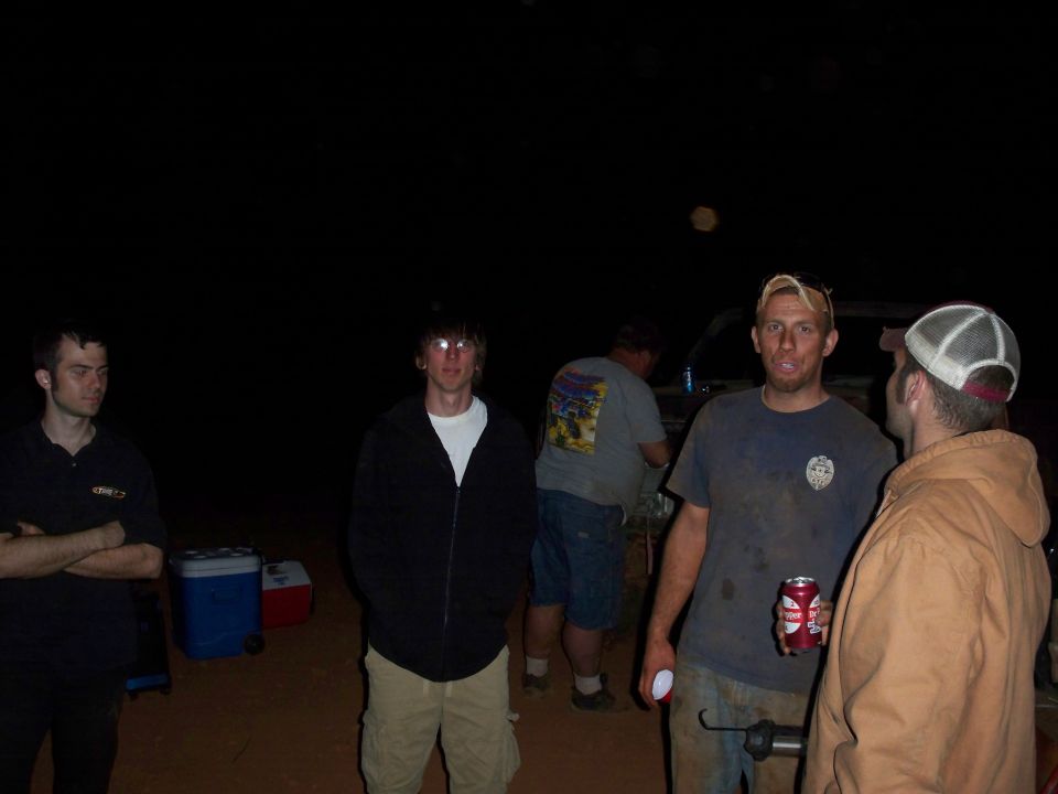 Four men stand around at night, one holding a can of beer