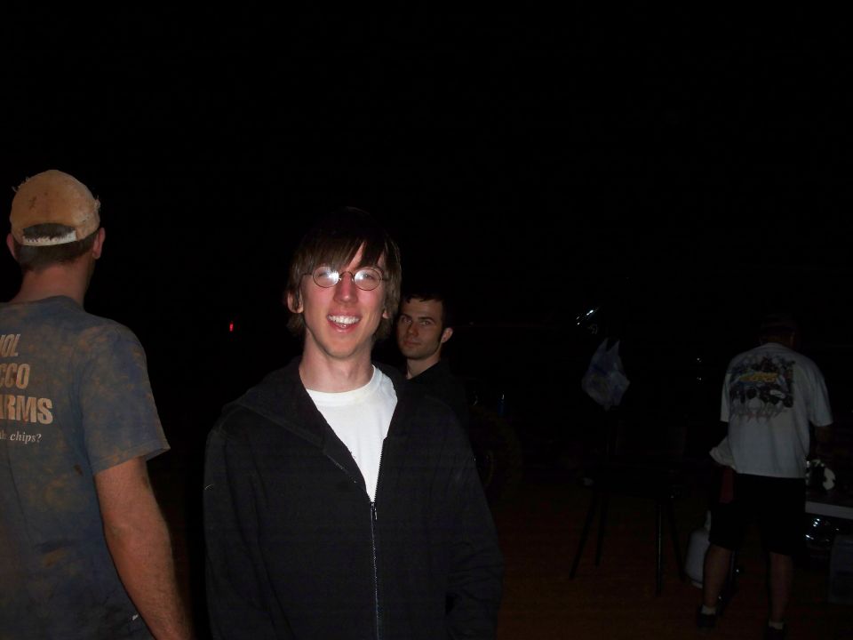 Three men stand in the dark, one in foreground wearing glasses and a black hoodie over a white shirt
