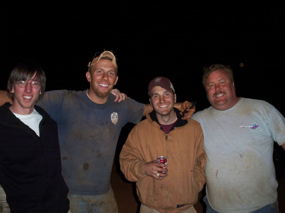 Four men, covered in dirt, pose for a photo at night