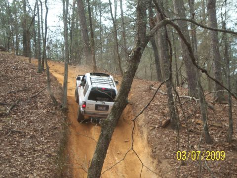 1996 Ford Bronco - Two SUVs navigate a challenging off-road trail through a dense forest on a cloudy day.