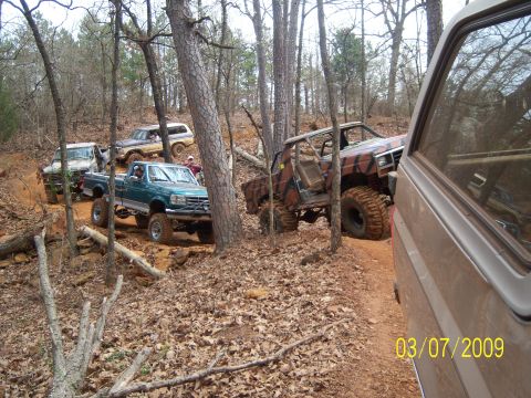 1996 Ford Bronco - Multiple off-road vehicles navigate a wooded trail on a sunny day.