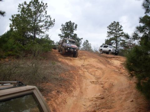 1996 Ford Bronco - Several off-road vehicles navigate a challenging dirt trail, ascending a steep incline under an overcast sky.