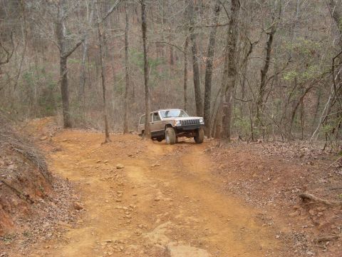 1996 Ford Bronco - A tan Jeep Cherokee XJ navigates a rutted dirt path through a forest.