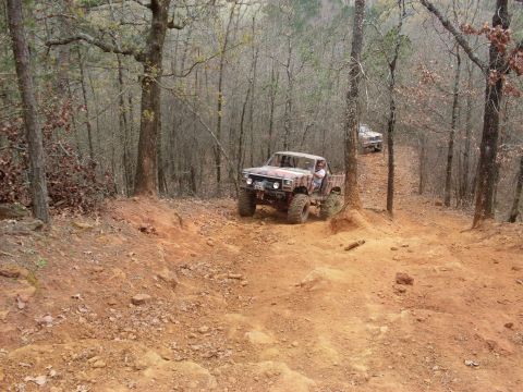 1996 Ford Bronco - Two off-road pickup trucks navigate a challenging, wooded trail with steep inclines and loose dirt.
